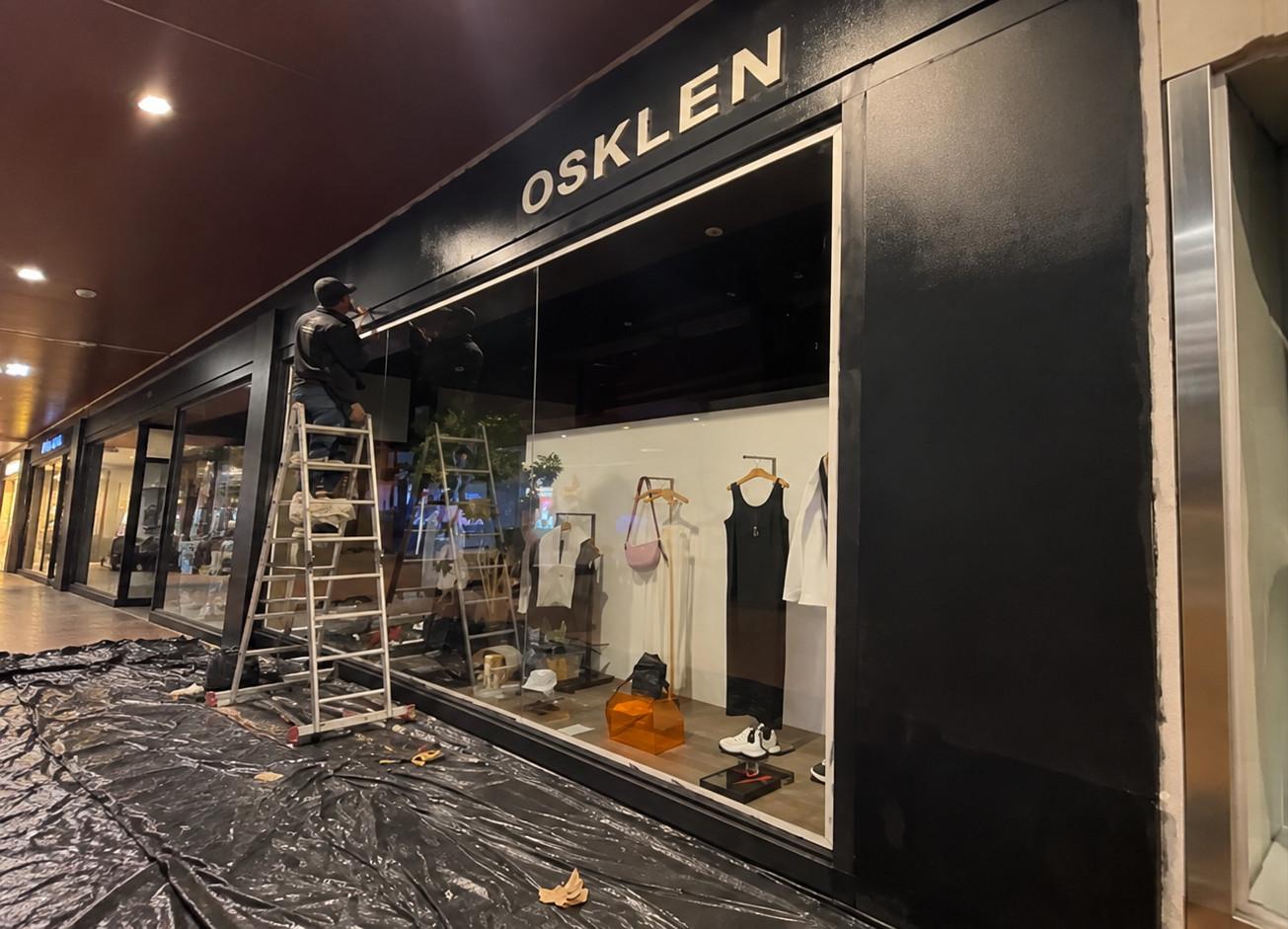 Profissional em escada pintando a fachada da loja Osklen em um shopping, com o piso protegido por lona