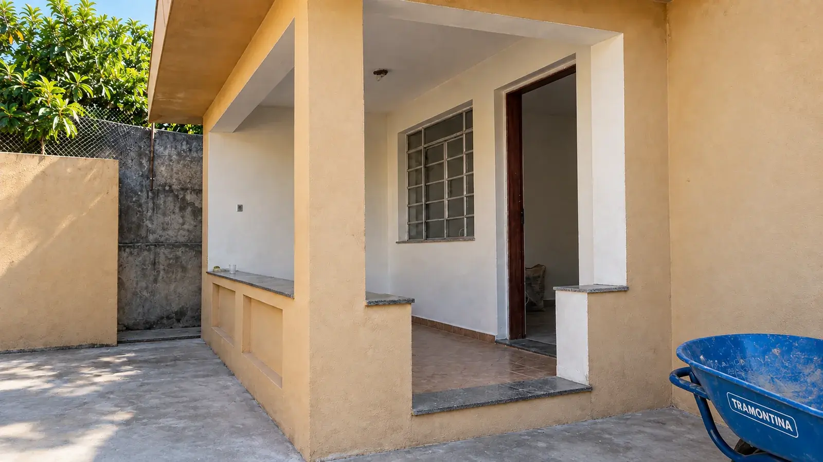 Varanda de casa renovada, com paredes pintadas, piso cerâmico e um carrinho de mão ao lado